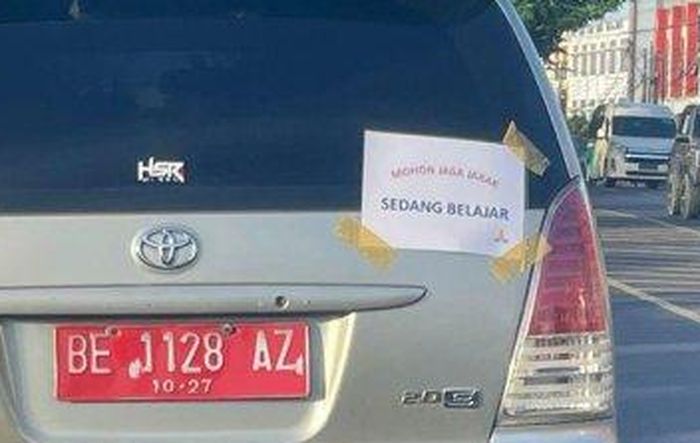 Tulisan di kertas yang menempel di kaca belakang Toyota Kijang Innova pelat merah BE 1128 AZ milik Pemkot Bandar Lampung bertuliskan 'MOHON JAGA JARAK, SEDANG BELAJAR'